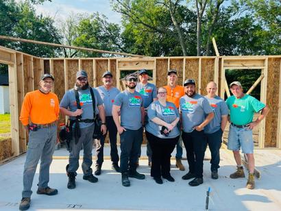 Ten people in construction gear pose and smile inside a partially built wooden structure on a sunny day.