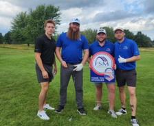 Four men standing on a golf course, one holding a circular FSM sponsor sign, all posing and smiling.