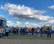 A crowd of people, including kids in sports uniforms, gathers near a food truck under a blue sky with clouds.