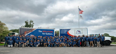 Large group of workers in blue shirts posing in front of company trucks and a flagpole under a cloudy sky.