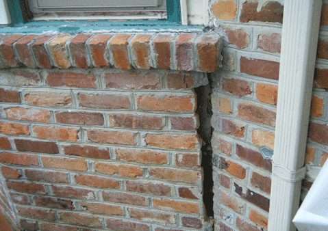 Large vertical crack in a brick wall near a window and drainpipe, indicating structural damage from foundation settlement.