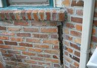 Large vertical crack in a brick wall near a window and drainpipe, indicating structural damage from foundation settlement.