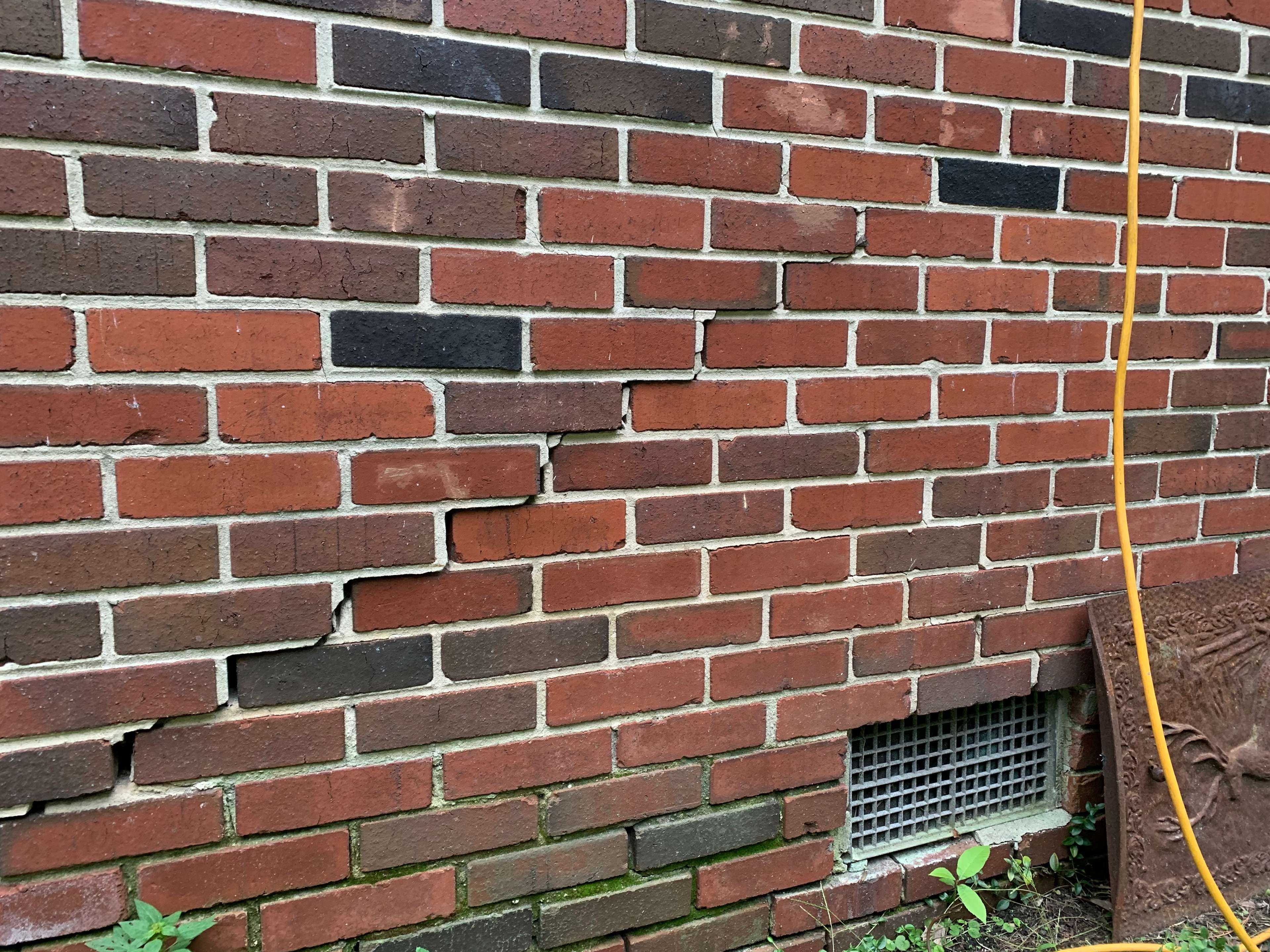 Cracked red brick wall with misaligned bricks above a vent and some grass at the bottom.