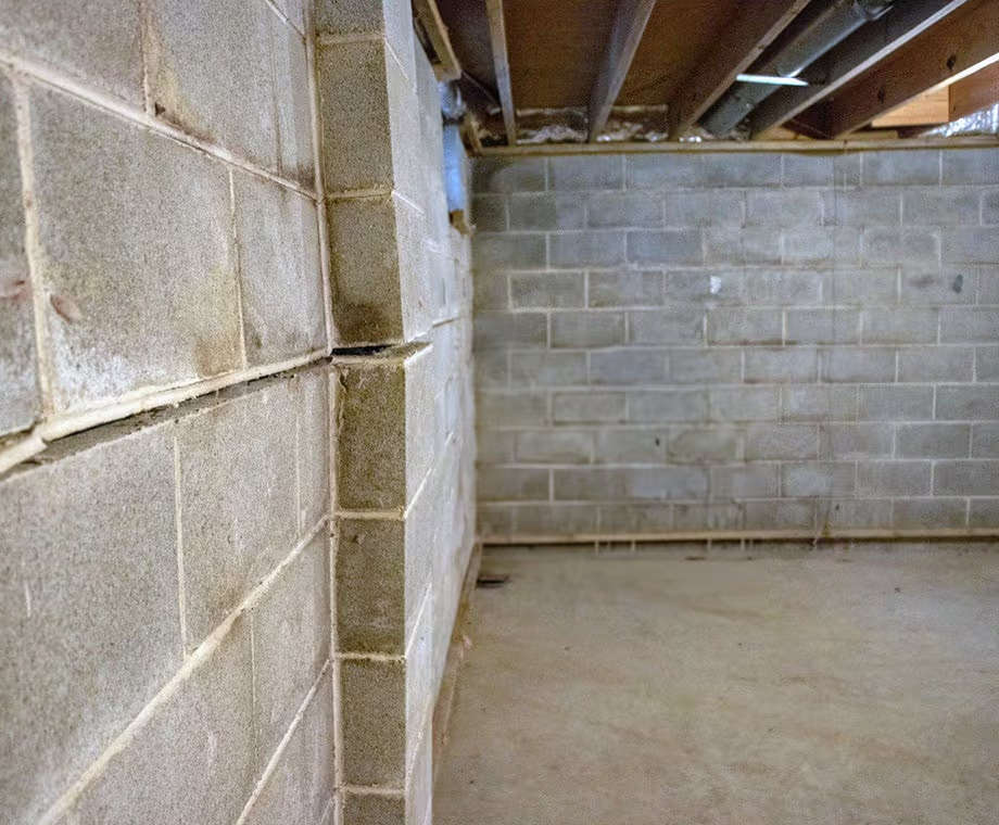 Cracked and bowed cinder block basement wall with exposed ceiling joists and concrete floor.