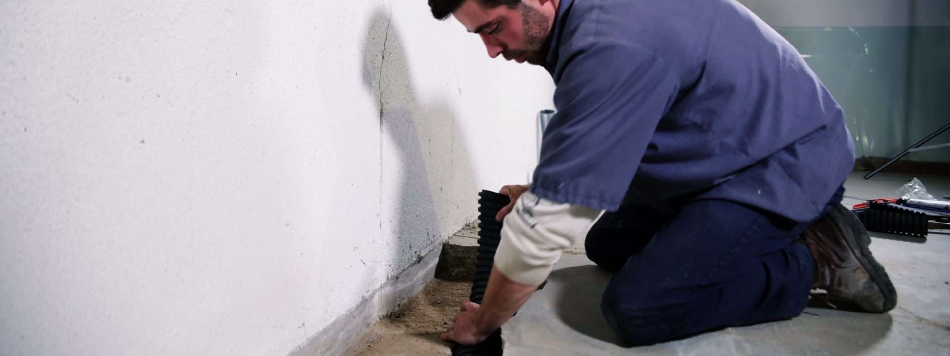 A man kneels, expertly installing waterproofing material along a basement wall, guarding against the 5 ways water enters your basement, ensuring a dry and secure foundation.