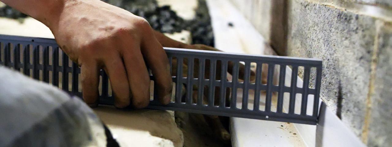 Hand installing a gray ventilated cover on a building surface to combat one of the 5 ways water enters your basement.