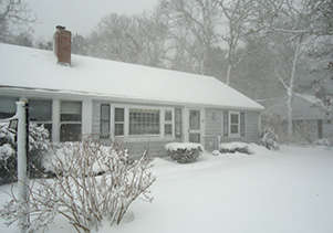 A house and yard covered in heavy snow during a winter snowstorm with trees in the background.
