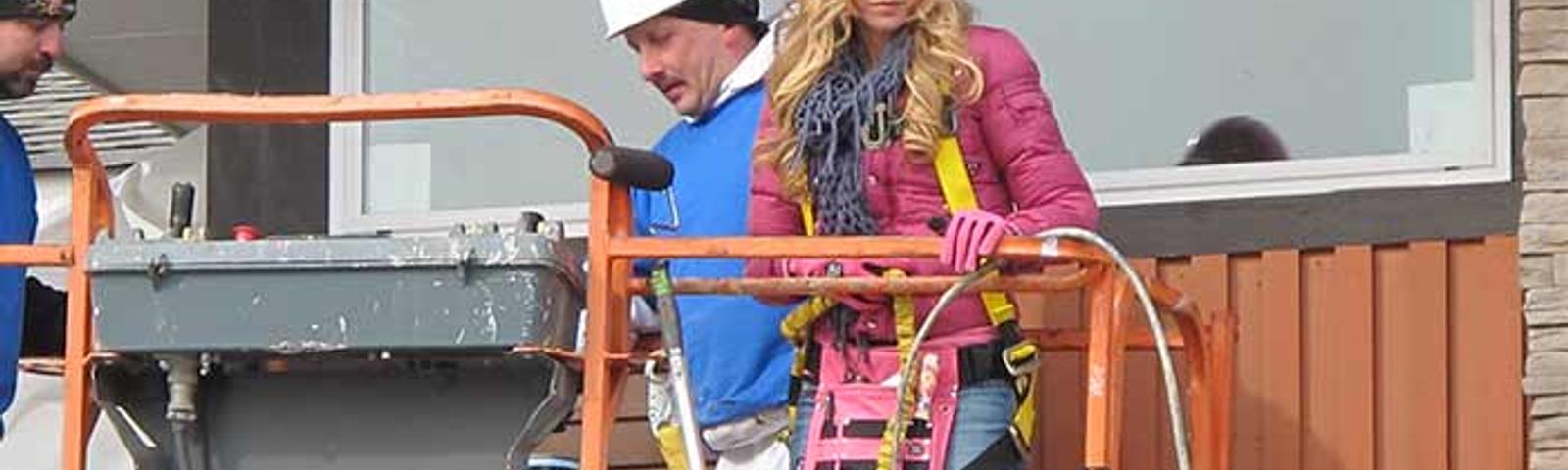 Two construction workers on a lift: one in a white hard hat and another in a pink hard hat, wearing safety gear.