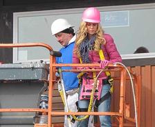 Two construction workers on a lift: one in a white hard hat and another in a pink hard hat, wearing safety gear.