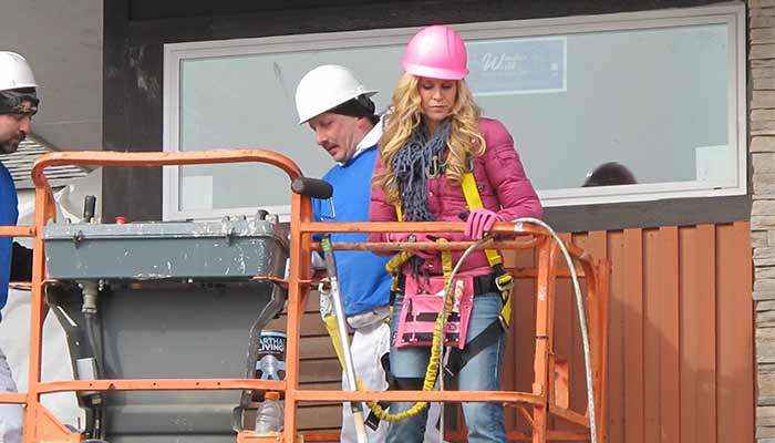 Two construction workers on a lift: one in a white hard hat and another in a pink hard hat, wearing safety gear.