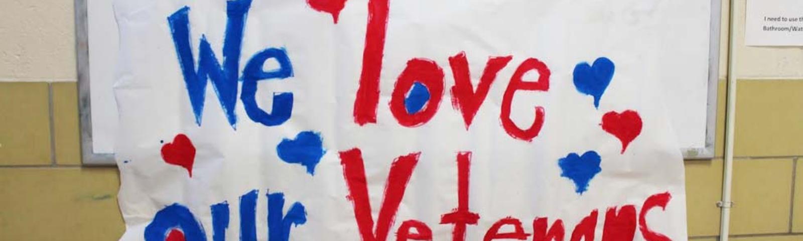 Handmade sign with red and blue letters reads "We love our Veterans," with hearts surrounding the text, beautifully capturing our heartfelt Veterans Day tribute in Norfolk, VA in 2015.