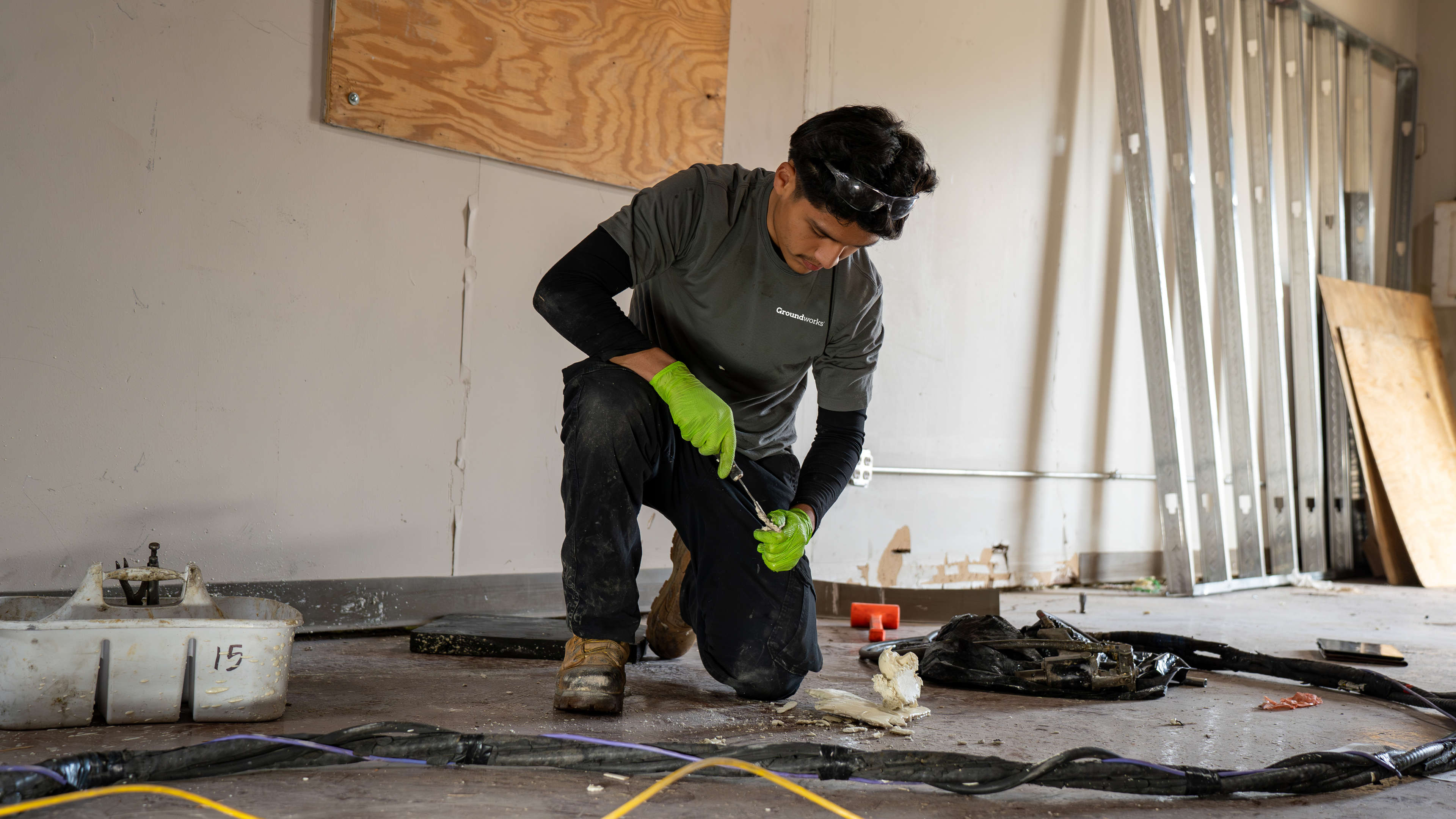 A person in gloves kneels on the floor, working with tools in a room under construction or renovation.