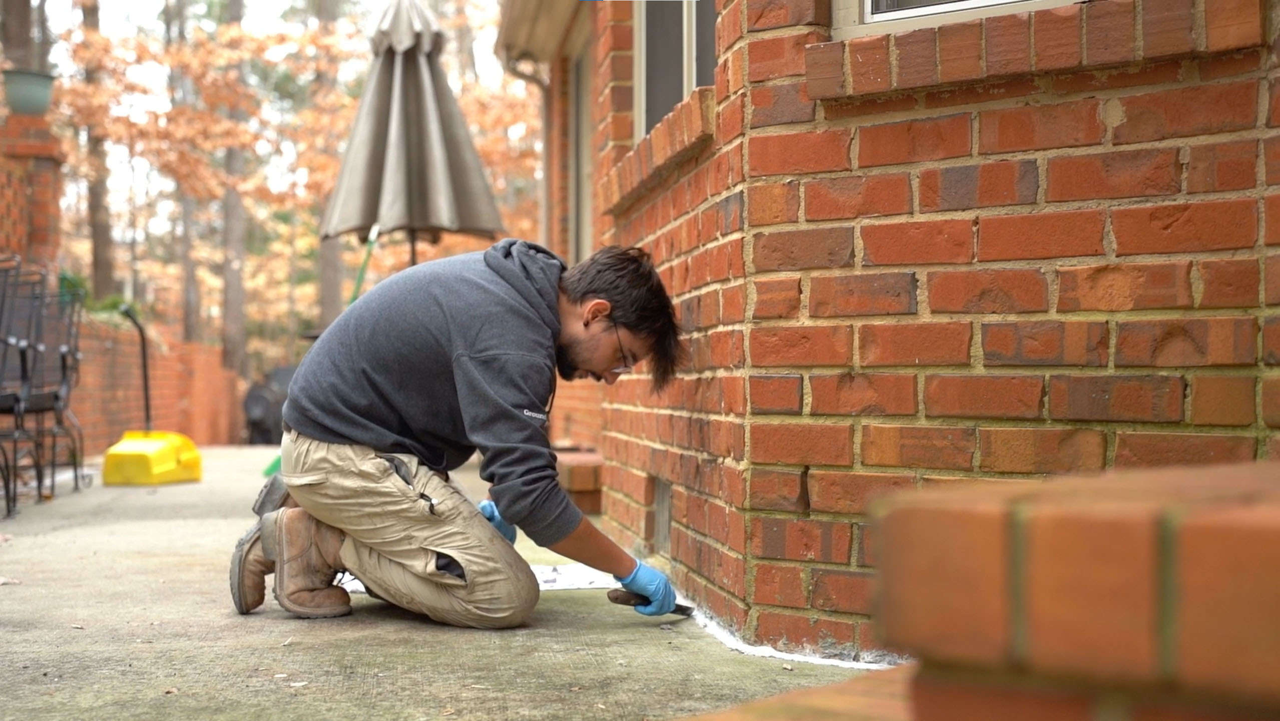 Person kneeling on concrete, applying sealant to the base of a brick wall with a caulking gun.