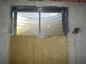 A basement window, partially covered by insulation, overlooks a steel window well with a hint of light suggesting the need for wet basement repair.