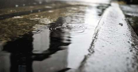 Raindrops falling into a puddle on a city street, with reflections visible on the wet surface.