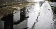 Raindrops falling into a puddle on a city street, with reflections visible on the wet surface.