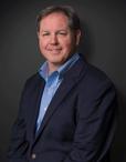 Steve Frey, recently appointed by JES, stands confidently in a dark suit and blue shirt, smiling warmly against a dark background.