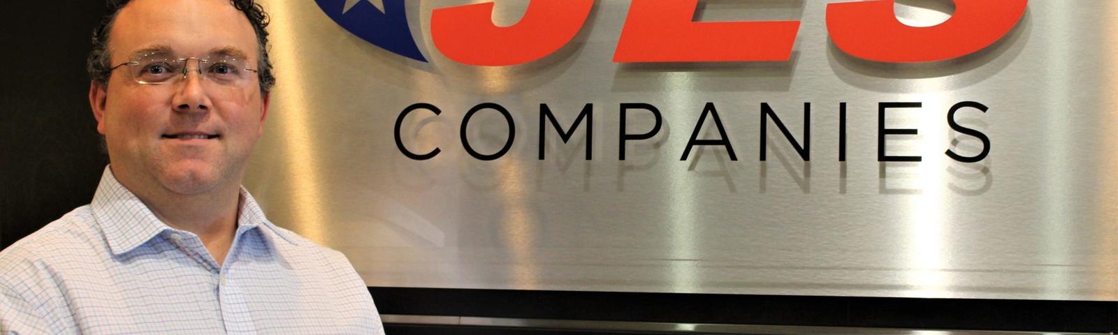 A man in a white shirt stands next to a JES Companies sign with a blue and red logo.