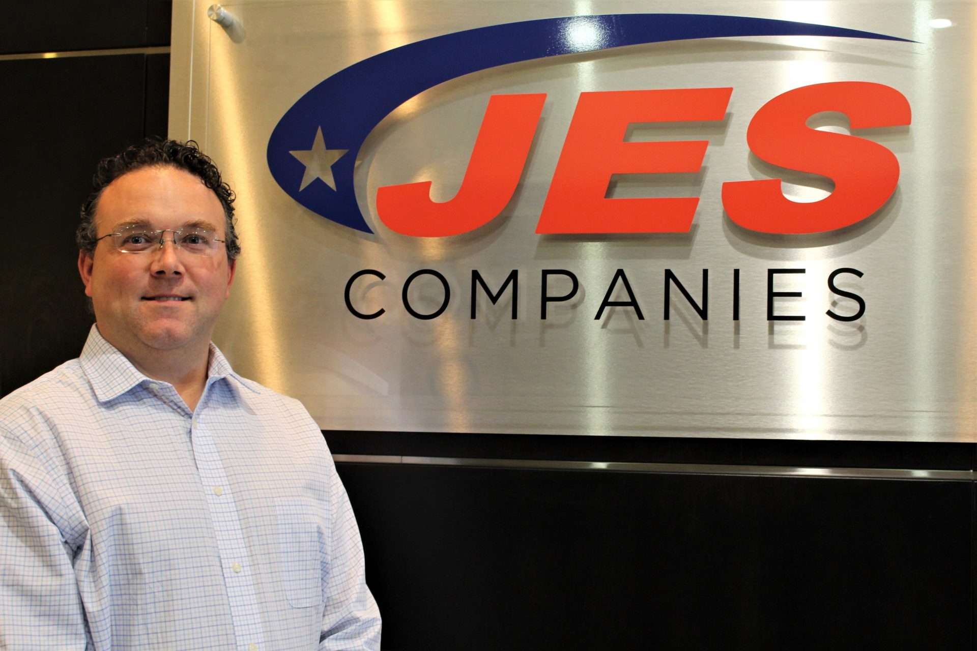 A man in a white shirt stands next to a JES Companies sign with a blue and red logo.