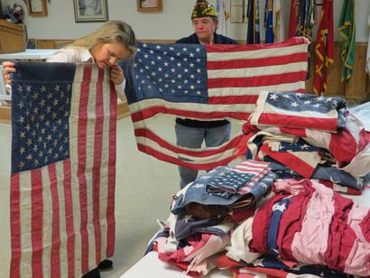Two people inspect worn American flags next to a table with a pile of folded flags in a room decorated with flags.