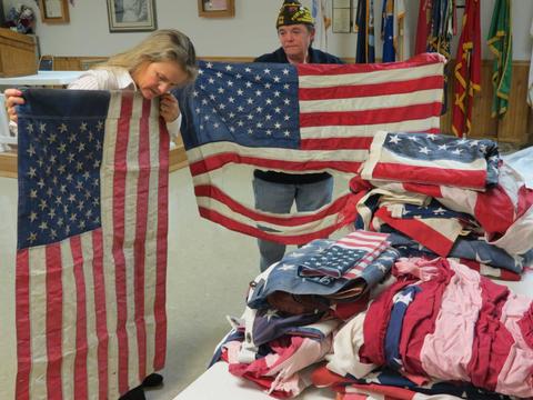 Two people inspect worn American flags next to a table with a pile of folded flags in a room decorated with flags.