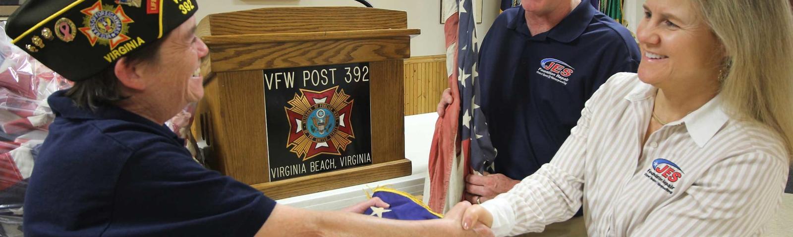 A veteran shaking hands with two people at a VFW post, holding an American flag.