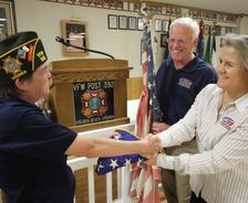 A veteran shaking hands with two people at a VFW post, holding an American flag.