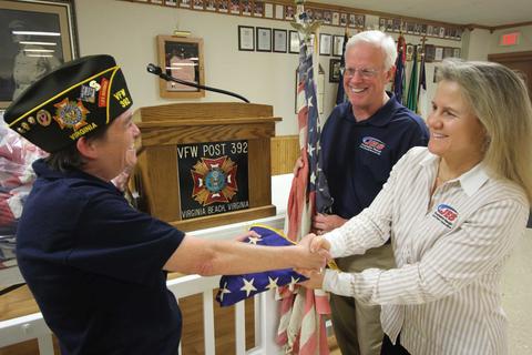 A veteran shaking hands with two people at a VFW post, holding an American flag.