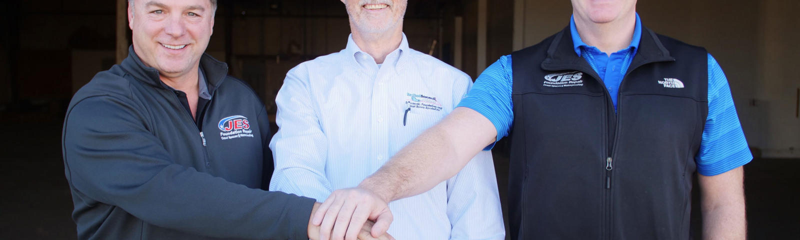 Three men smiling, standing arm in arm in a warehouse, wearing casual business attire.