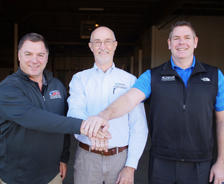Three men smiling, standing arm in arm in a warehouse, wearing casual business attire.