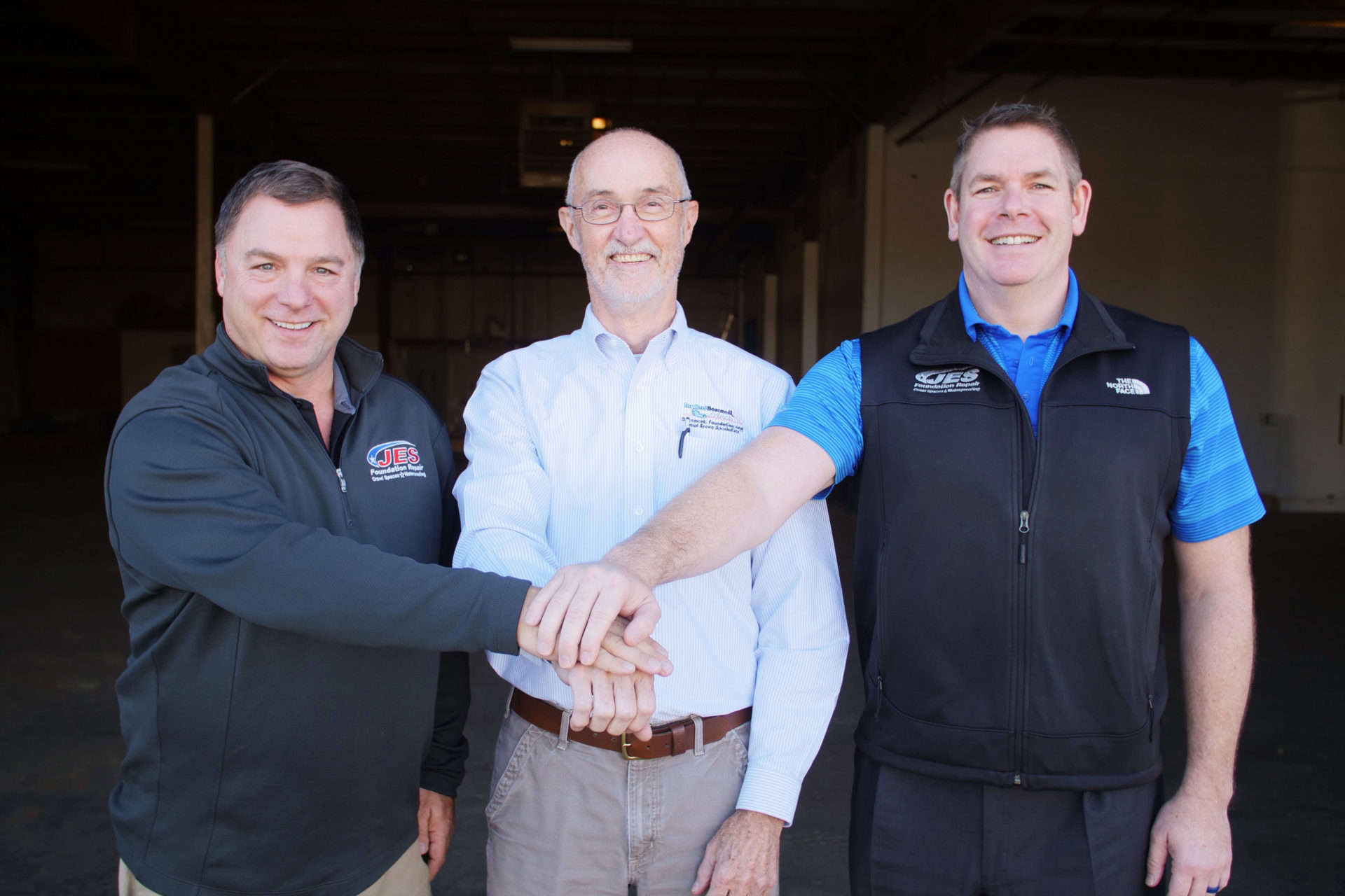 Three men smiling, standing arm in arm in a warehouse, wearing casual business attire.