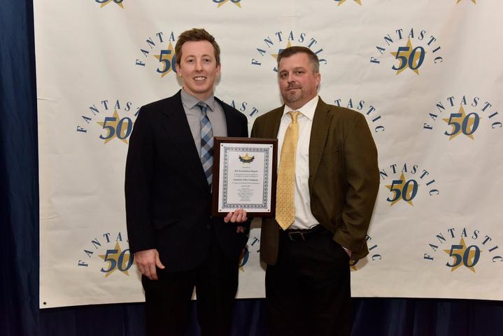 Two men in suits stand in front of a “Fantastic 50” backdrop, one holding a framed certificate.