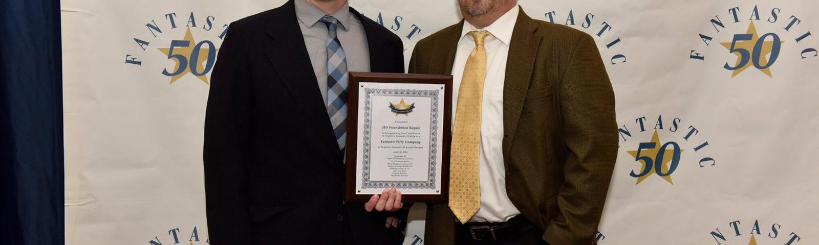Two men in suits stand in front of a “Fantastic 50” backdrop, one holding a framed certificate.