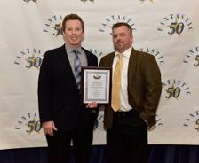 Two men in suits stand in front of a “Fantastic 50” backdrop, one holding a framed certificate.