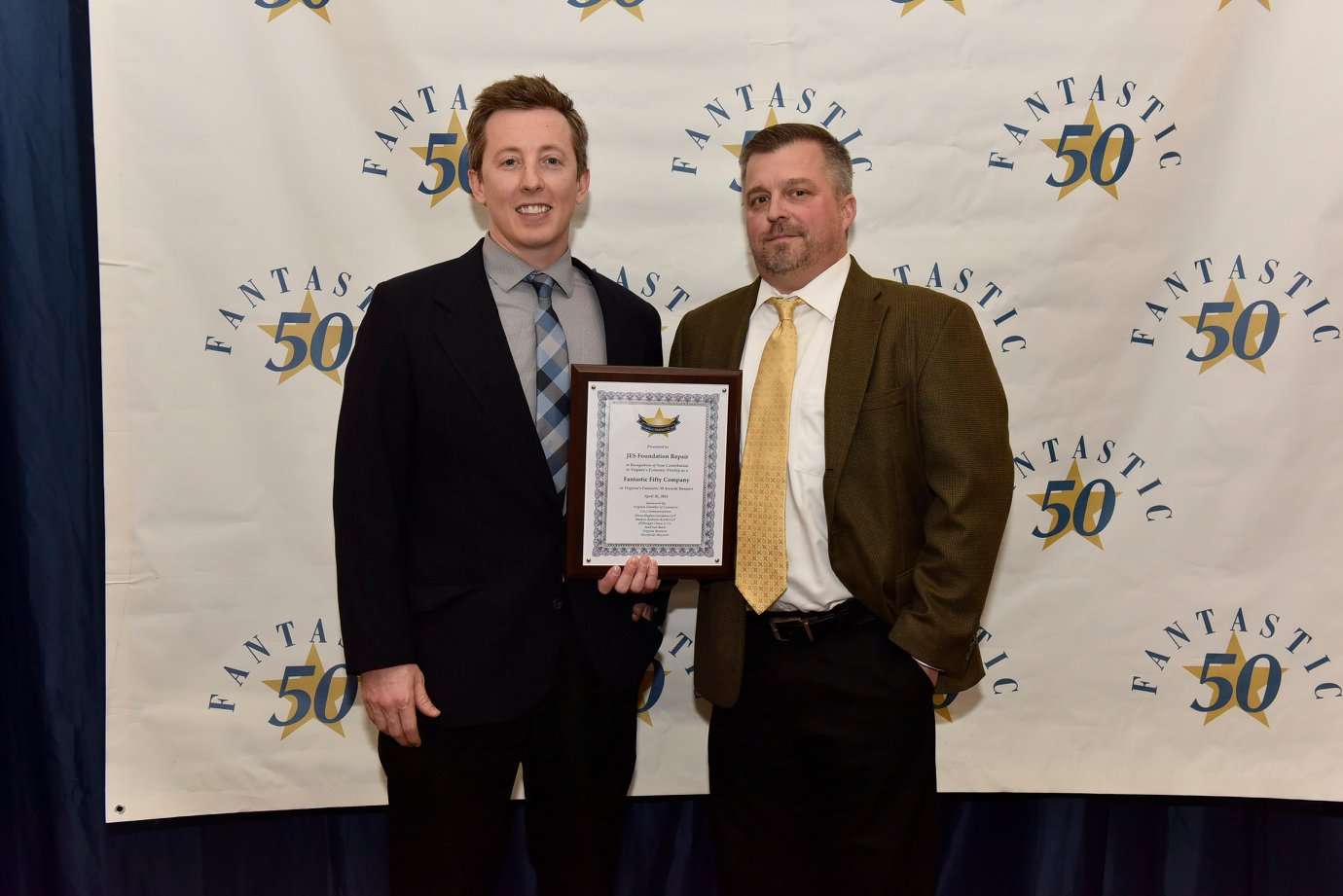 Two men in suits stand in front of a “Fantastic 50” backdrop, one holding a framed certificate.