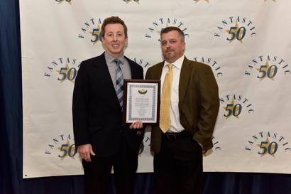 Two men in suits stand in front of a “Fantastic 50” backdrop, one holding a framed certificate.