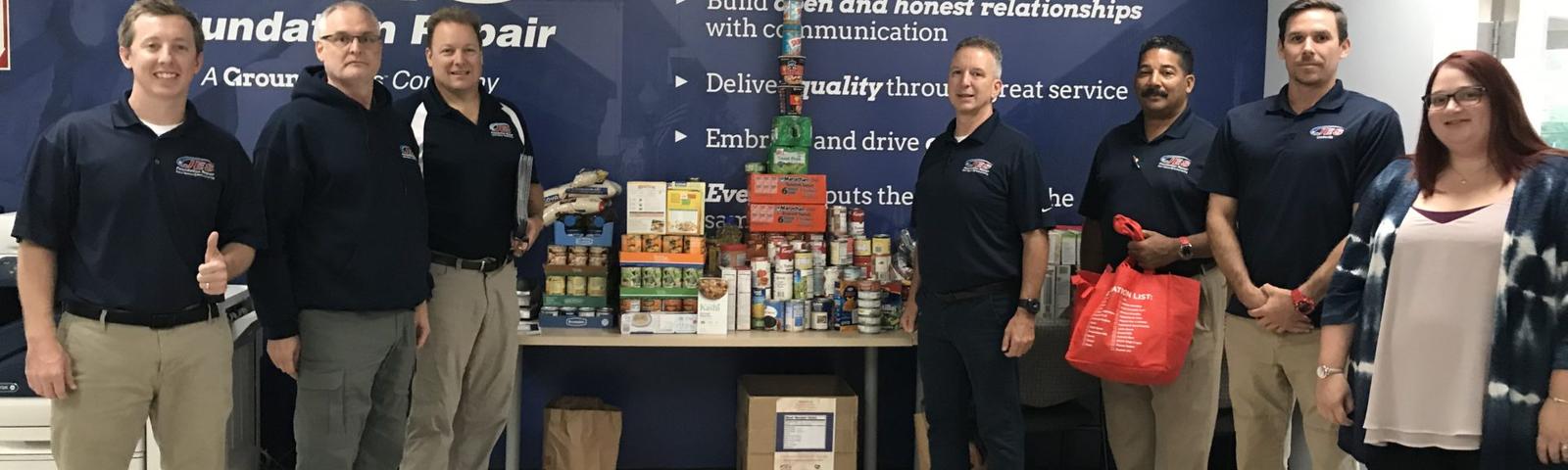 A group of people stands by donated food items, supporting the hungry, in front of a "JES Foundation Repair" sign and a core values wall display.