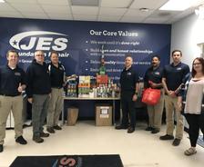 A group of people stands by donated food items, supporting the hungry, in front of a "JES Foundation Repair" sign and a core values wall display.