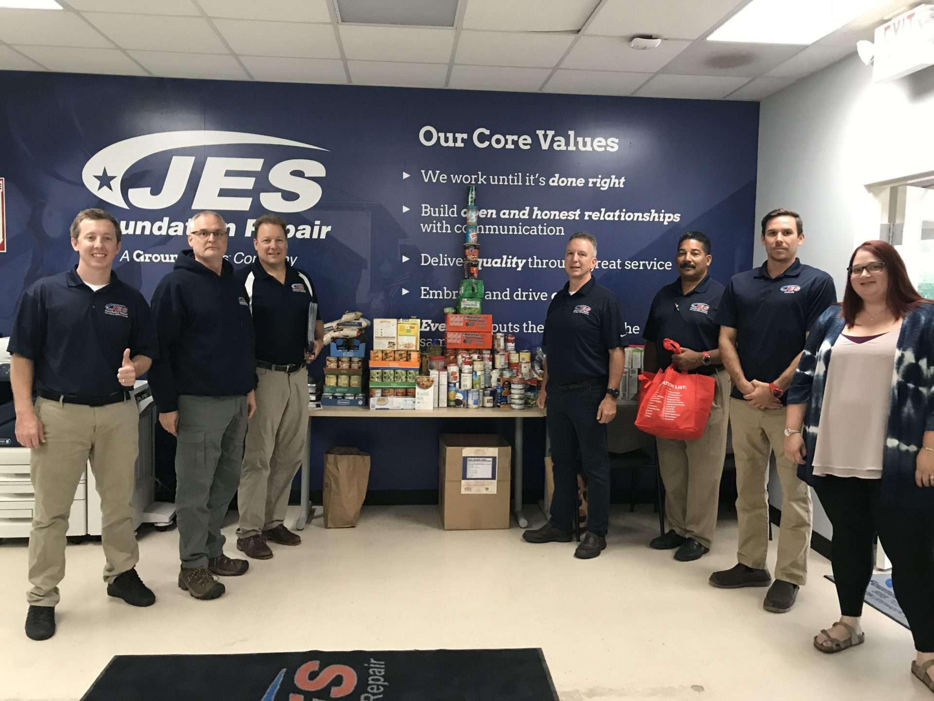 A group of people stands by donated food items, supporting the hungry, in front of a "JES Foundation Repair" sign and a core values wall display.