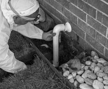 Person in protective suit inspecting a pipe near a brick wall, surrounded by grass and rocks.
