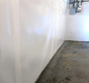 A basement wall with white plastic sheeting and a bare concrete floor, partially covered near the wall.