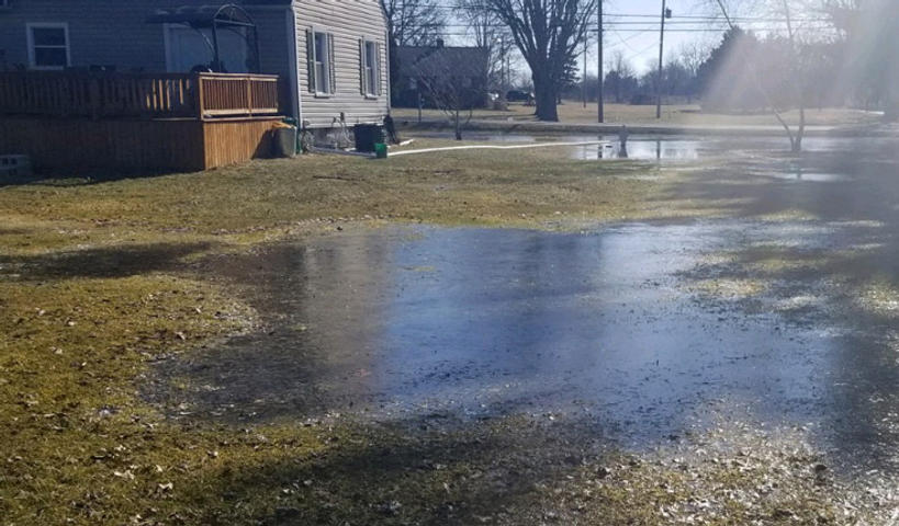 Large puddle of water covering a grassy yard near a house on a sunny day.