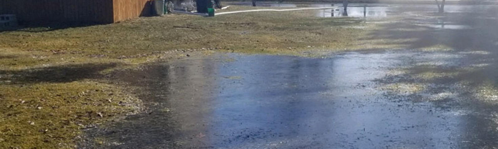 Large puddle of water covering a grassy yard near a house on a sunny day.