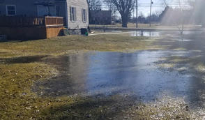 Large puddle of water covering a grassy yard near a house on a sunny day.