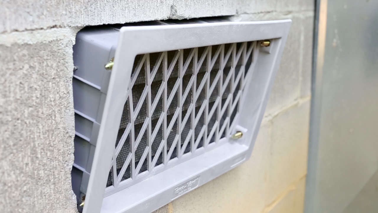 A close-up of a gray plastic crawlspace vent installed in a concrete wall.