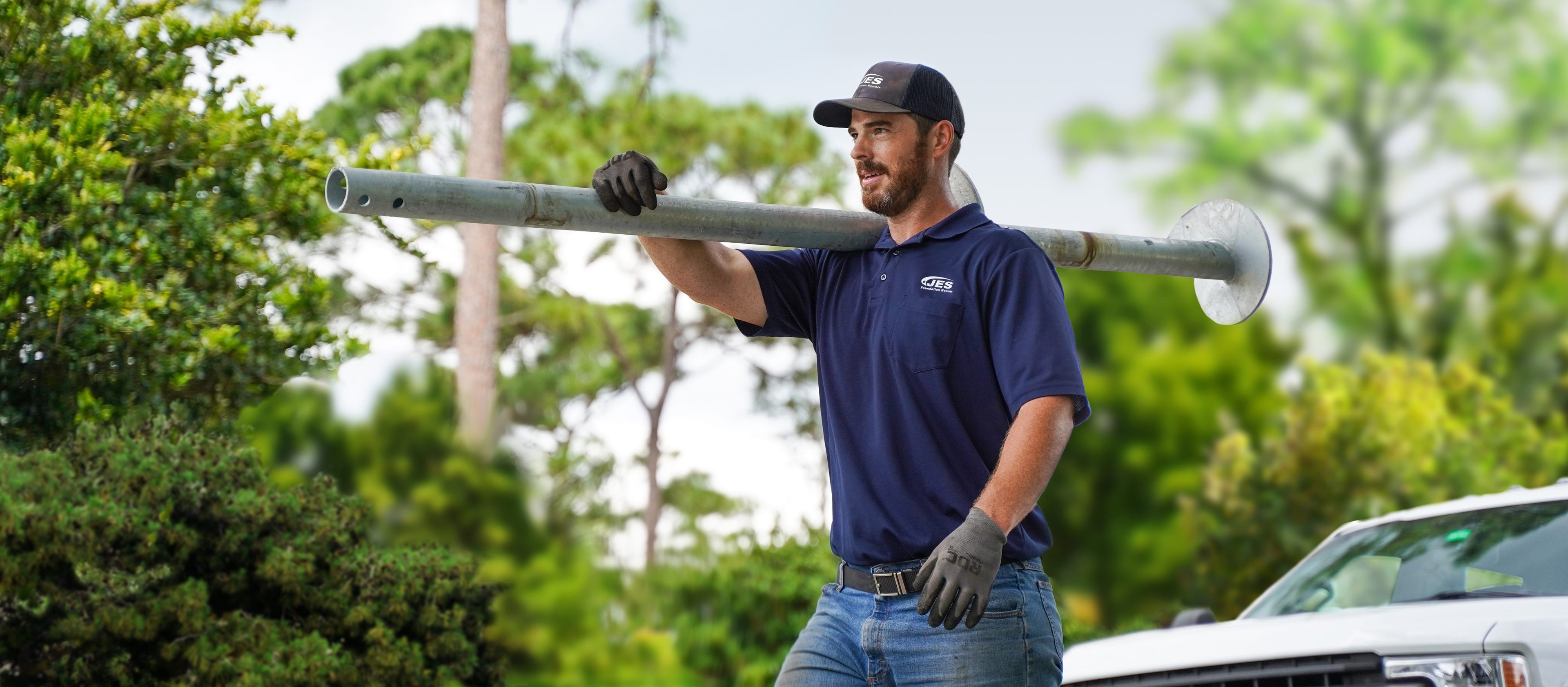 A man in work gloves carries a foundation pier on his shoulder outdoors near trees and a white truck.