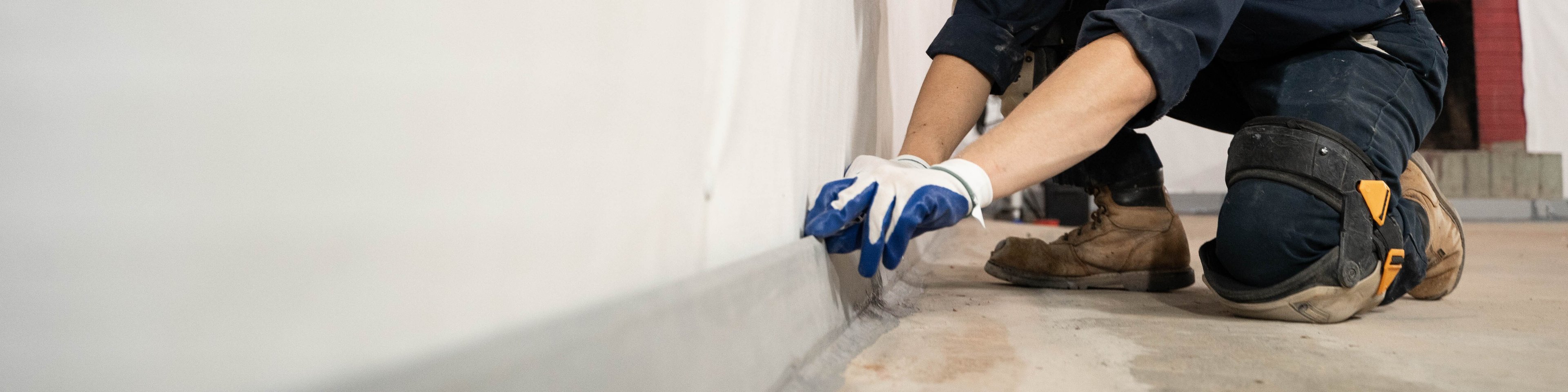 A worker in gloves and a cap installs or inspects baseboard trim along a white wall in a construction area.