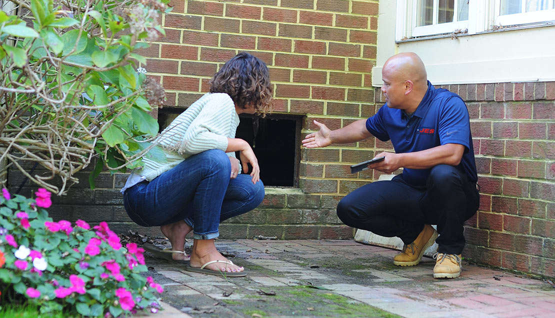 Two people are examining a brick wall opening outside a building, where the subtle scent of flowers mingles with the cool air from basement condensation.