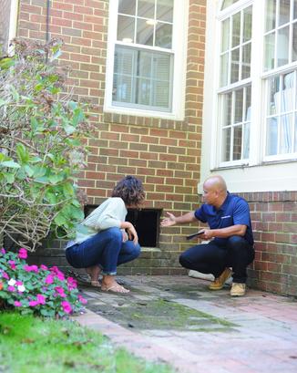 Two people crouch by a brick house's foundation, discussing repairs near the open crawl space vents, with flowers nearby.
