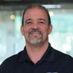 A man with a goatee smiles, wearing a dark polo shirt with a "JES" logo. Blurred background with soft lighting.